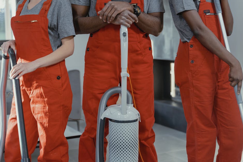 Two professional cleaners from Islington Cleaners, wearing beige uniforms with white collars, stand side by side against a plain white wall in a well-lit indoor setting. The cleaner on the left holds 