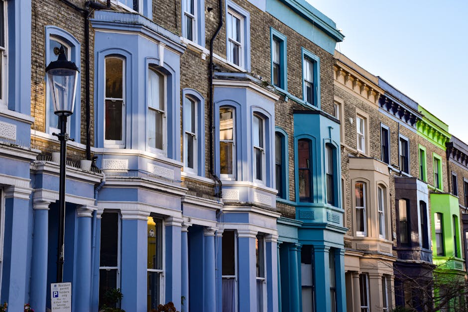A row of Victorian-style terraced houses with colorful facades in shades of blue, beige, and green, featuring large bay windows with white frames, decorative brickwork, and ornate cornices. The building surfaces are clean and well-maintained, with glass on the windows reflecting the sky. A traditional black street lamp stands in front, and the scene is lit by natural daylight, highlighting the neat appearance of the exteriors. The image showcases residential architecture typical of Islington, emphasizing cleanliness and the quality of surface finishes, associated with professional domestic cleaning services provided by Islington Cleaners. This vibrant and tidy street scene underscores the importance of careful surface cleaning and maintenance in urban residential areas.