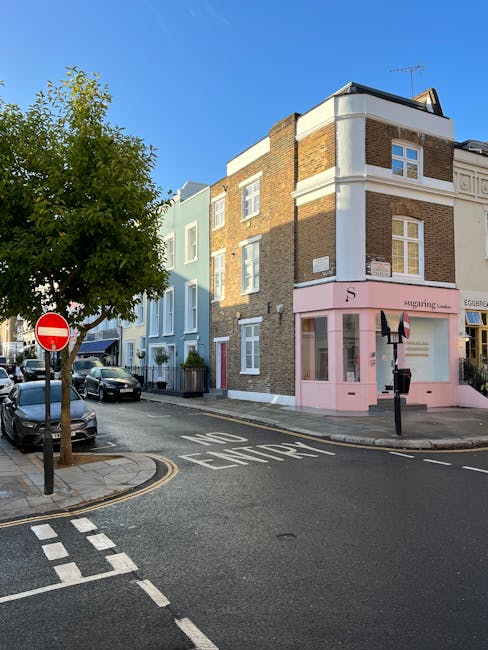 Image of a corner building in Islington with a pastel pink shopfront labeled 'S', featuring large display windows and a pastel pink door. The building has a white curved architectural detail on the upper floor and brickwork on the lower floors. Surrounding the building are parked cars along the street, a tree with green foliage on the left, and a clear blue sky overhead. The adjacent street has a 'NO ENTRY' traffic sign and marked road lines, indicating a one-way street. The scene reflects a quiet urban environment with maintained roads and streetscape, typical of residential and commercial areas in Islington. This image emphasizes the clean and well-kept appearance of the area, aligning with professional cleaning standards provided by Islington Cleaners.