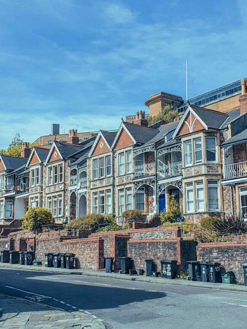 A row of Victorian-style terraced houses with colorful facades in shades of blue, beige, and green, featuring large bay windows with white frames, decorative brickwork, and ornate cornices. The building surfaces are clean and well-maintained, with glass on the windows reflecting the sky. A traditional black street lamp stands in front, and the scene is lit by natural daylight, highlighting the neat appearance of the exteriors. The image showcases residential architecture typical of Islington, emphasizing cleanliness and the quality of surface finishes, associated with professional domestic cleaning services provided by Islington Cleaners. This vibrant and tidy street scene underscores the importance of careful surface cleaning and maintenance in urban residential areas.
